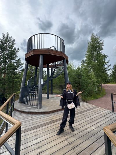 A woman in black athletic wear and a black baseball cap stands on a wooden observation deck in front of a spiral staircase leading to a multi-tiered observation tower. She is facing the camera with her arms outstretched and a small, patterned crossbody bag hanging from her shoulder. The tower, constructed with dark wood and metal railings, is set within a forest of pine and birch trees under a cloudy sky. The location appears to be an outdoor recreational area.