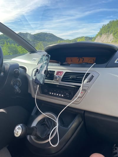 The image shows the interior of a car parked on a road in Očinići, Montenegro. The dashboard features a digital speedometer displaying "77" km/h, an air conditioning vent, and a car stereo. A smartphone is mounted on a holder on the dashboard, connected to a white charging cable. The car's gear shift, a 6-speed manual transmission, is visible in the foreground. The background reveals a sunlit, mountainous landscape with lush green vegetation. The sky is clear with scattered clouds.