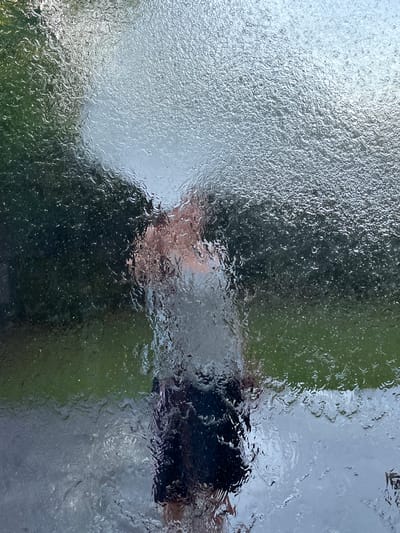 This image captures a distorted reflection of a person on a textured, wet surface, possibly glass or plastic with water droplets. The person appears to be wearing dark clothing and is seen from the chest up, with their face partially visible and heavily blurred. The background is a mix of green and gray, suggesting an outdoor environment with foliage and possibly a sky or building. The texture on the surface creates a rippling effect, making it difficult to discern fine details. Small, white specks are scattered across the surface, possibly representing rain or condensation. The overall impression is one of a cloudy or rainy day, given the wetness and muted light. The location is specified as Mondercange, Luxembourg. There is no visible text in the image.