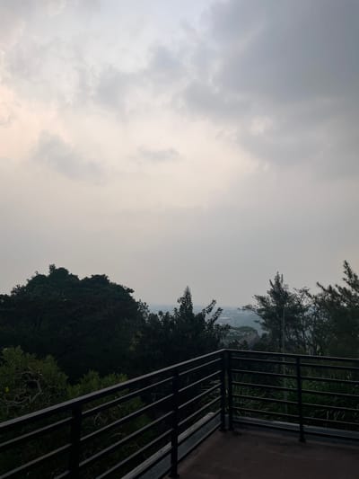 This is a photograph taken from a balcony overlooking a landscape in Babakan Madang, Indonesia. The sky is overcast with muted gray and white clouds. In the foreground, a dark metal railing runs horizontally across the lower portion of the image, with a concrete floor visible beneath it. Beyond the railing, a dense collection of trees and foliage dominates the middle ground. The trees are dark green, with some lighter green vegetation interspersed. In the distance, a hazy view of a city or town can be seen, with rooftops and buildings partially obscured by atmospheric conditions. The overall impression is one of a somewhat subdued and misty environment, typical of an early morning or late afternoon in a tropical region. No people are visible in the frame.