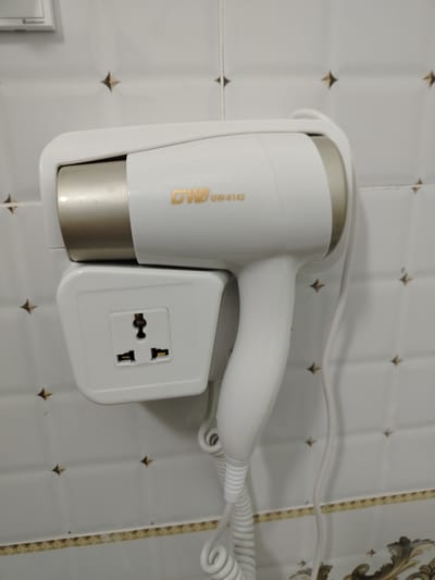 A white hairdryer with a gold accent is mounted on a wall. Below the hairdryer is a white wall-mounted electrical outlet with three different types of sockets. The wall appears to be tiled with a pattern of small, brown star-shaped tiles. The hairdryer has the model number "GW-6142" printed on it in gold lettering. The cord of the hairdryer is coiled and hangs down. The scene is well-lit, and the overall impression is of a hotel bathroom or similar establishment. The image does not provide any specific details about the city of Pitnak, Uzbekistan.