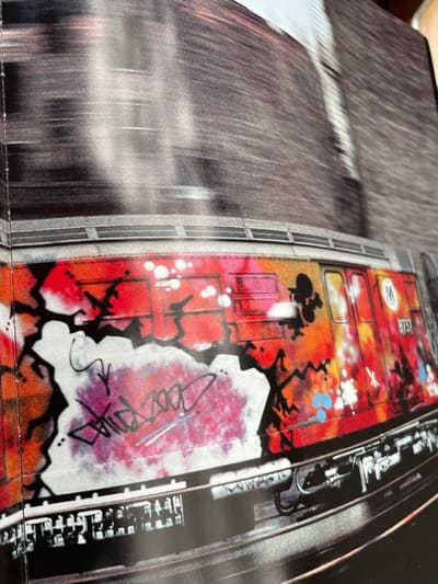 The image is a close-up of a red train car covered in graffiti. The train is located at the coordinates 44.882, -72.565, which is in the city of Montpelier, Vermont. The graffiti is in various colors, including white, black, blue, orange, and pink. The graffiti is mostly abstract, but there are also some legible words and symbols. The train is stationary and appears to be on tracks. There are no people visible in the image. The image is taken at an angle and the train is in focus, while the background is out of focus.
