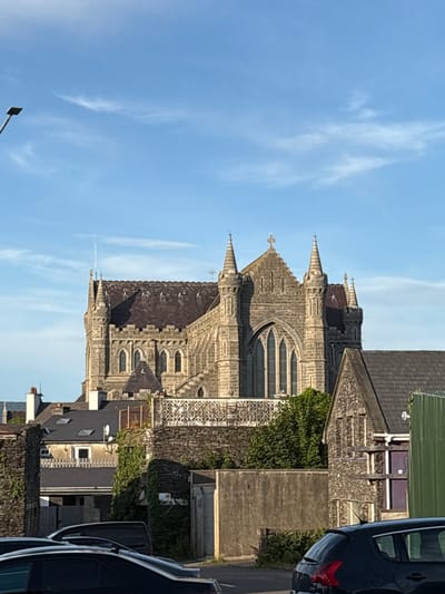 The image shows a stone church with a tall steeple against a blue sky with scattered clouds. In the foreground, there are several cars parked on a street, and some residential buildings and stone walls. The church appears to be a historical building with Gothic architectural features, such as arched windows and pointed spires. The overall scene depicts a town in daylight, with the church as the prominent landmark. The location is identified as Cahersiveen, Ireland.