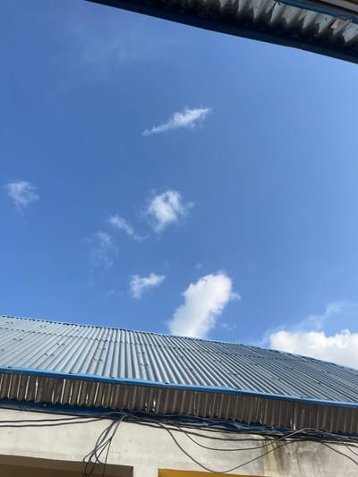 The media file shows a view of the sky from a low angle, looking upwards. The scene is dominated by a clear blue sky with scattered white clouds. On the right side of the frame, a corrugated metal building with a blue trim runs vertically. Electrical wires are visible along the side of an adjacent wall, which appears to be painted in a light color, possibly concrete. On the left side of the frame, a metal structure, possibly part of a roof or overhang, is visible with a dark blue trim. The lighting suggests it is daytime, with bright sunlight illuminating the scene and casting shadows. The clouds are wispy and appear to be thin, indicating fair weather. There are no people or any clear indication of a specific activity taking place. The setting appears to be an urban or semi-urban environment, given the presence of the building and electrical infrastructure. There is no visible text in the image.