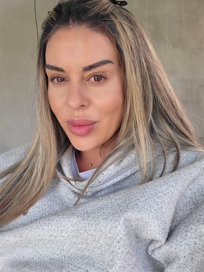 A medium-shot, close-up selfie shows a woman with long, blonde and brown ombre hair pulled back partially with a black clip. She has brown eyes, dark eyebrows, and full lips with a light pink lipstick. She is wearing a light grey hooded sweatshirt over a white t-shirt and a thin silver necklace with a small pendant. The background is a plain, light beige concrete wall. The image was taken outdoors in Ravnogor, Bulgaria.