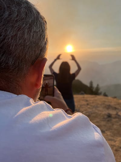 In the foreground, the back of a man's head and shoulder are visible, with his gray hair catching the golden light of the sunset. He is holding a smartphone in his right hand, angled towards a woman in the mid-ground. The woman is silhouetted against the bright sun, which she appears to be framing with her raised hands. She stands on a grassy, uneven terrain with distant mountains and trees visible in the background. The scene takes place outdoors during what appears to be sunset.