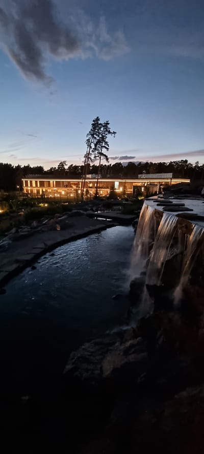 The image depicts a building with illuminated windows, suggesting it is evening or night. The building appears to be multi-storied and is surrounded by trees. In the foreground, there is a body of water with a waterfall cascading into it. The sky is a deep blue, transitioning to a lighter hue towards the horizon, indicating dusk or twilight. There are some clouds present. The scene suggests an outdoor setting, possibly a resort or a recreational area, given the presence of water features and a well-lit building. The water is dark and reflects the artificial lights from the building. The overall mood is tranquil and serene. No visible text is present in the image.