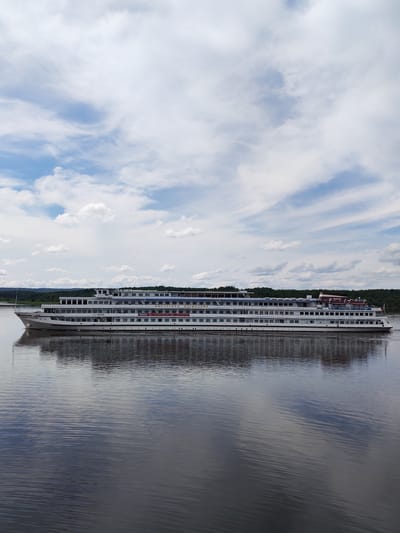 A wide, modern passenger ship with multiple decks is shown sailing on a calm river under a cloudy sky. The ship is white with blue and red accents and displays Russian text on its side. Its reflection is visible on the dark water. The background features a lush green shoreline with trees and rolling hills, indicative of a rural setting near Shchetinino, Russia. The sky is partly cloudy with dramatic white clouds against a bright blue sky.