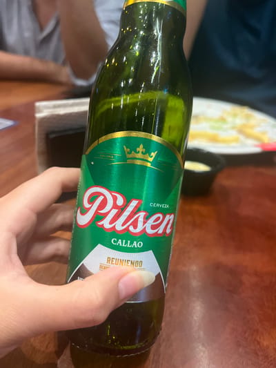 A person's hand holds a green glass bottle of Pilsen Callao beer. The label on the bottle is predominantly green with gold accents. It features the Pilsen logo in red and white, the word "CERVEZA" in white, and "CALLAO" in white. Below this, the word "REUNIENDO" is visible in white text on a dotted background. The background suggests an indoor setting with a wooden table, and out-of-focus individuals in the background. The image is a close-up of the beer bottle and the hand, with no discernible landmarks or specific details that would indicate the location within Santiago de Surco, Peru.