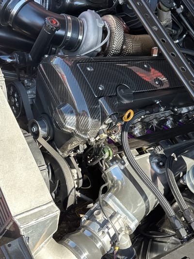 The image shows a high-performance car engine bay. Prominent features include a carbon fiber valve cover, a turbocharger with heat wrap, a billet aluminum intake manifold, fuel injectors with purple anodizing, and various braided stainless steel hoses and lines. The engine is belt-driven, with visible pulleys and a serpentine belt. An intercooler is visible at the bottom of the image. There is no indication of location or presence of any people.