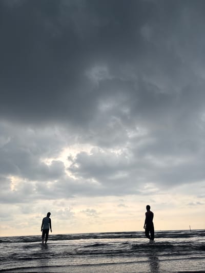 The image captures a coastal scene, likely a beach, under a dramatic sky filled with storm clouds. Two silhouetted figures are visible along the shoreline. One figure stands in the water near the top edge of the frame, while the second is positioned lower down. The environment consists of a dark, turbulent sea meeting a sky characterized by varying cloud formations. The location appears to be a beach in Angkleng Barat, Indonesia.