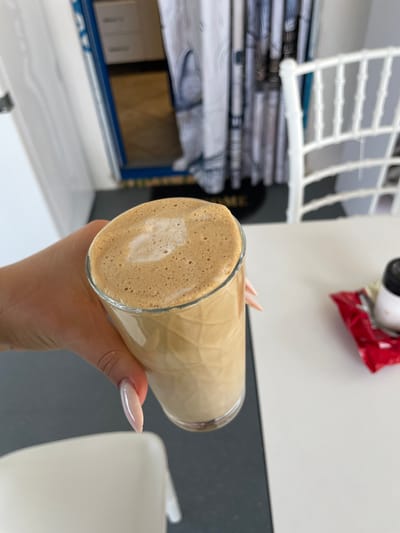 A hand with long, almond-shaped nails, painted in a light pink hue, holds a tall, clear glass filled with a frothy, light brown beverage, likely coffee or a similar drink with milk foam. The foam on top of the drink has a subtle heart-shaped pattern. The setting appears to be a cafe or a dining area, with a white table in the foreground and a dark grey surface to the left. In the background, a white chair with a distinctive design is visible, along with a white curtain and what looks like part of a doorway with blue trim, potentially leading to another room. A red packet, possibly containing sugar or a condiment, is on the table to the right, next to a small white object with a black lid. The scene is indoors, with diffused lighting. The location is identified as Skala Avdiron, Greece.