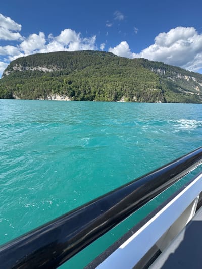 The image captures a view from a boat on a bright, sunny day, likely on Lake Thun, near Sigriswil, Switzerland. The turquoise water is rippling, with a wake trailing behind the unseen boat. In the background, a large, forested mountain dominates the horizon, its slopes covered in dense green trees. A rugged, rocky cliff face is visible partway up the mountain. The sky is a vibrant blue, dotted with fluffy white clouds. A black and white railing from the boat is prominently featured in the foreground, partially obscuring the view of the water.