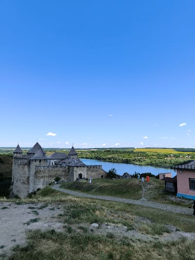 This image captures an elevated, wide-angle view of the Khotyn Fortress, situated on the right bank of the Dniester River in Khotyn, Chernivtsi Oblast, Ukraine, under a clear blue sky. The fortress, constructed from grey stone, occupies the left side of the frame, featuring multiple conical-roofed towers and high curtain walls, with an arched entrance visible in its central section.

The Dniester River, a wide body of calm blue water, extends horizontally behind the fortress towards the right. Beyond the river, the landscape consists of dense green foliage, with a distant yellow field, likely sunflowers, visible on a slight rise.

The foreground and midground are composed of an undulating grassy hillside traversed by a winding path of packed earth and uneven flagstones. Patches of dry earth and sparse vegetation are present across the ground. Several individuals are visible in the midground: two near the fortress entrance, one wearing a long white garment; and two further right, near the riverbank, one distinctly clad in a bright orange/red full-length garment. On the far right, a light pink building with a dark roof and dark-framed windows is partially visible, with a dark-clad individual partially visible adjacent to it. The scene is illuminated by natural daylight, indicated by the clear sky and subtle shadows.