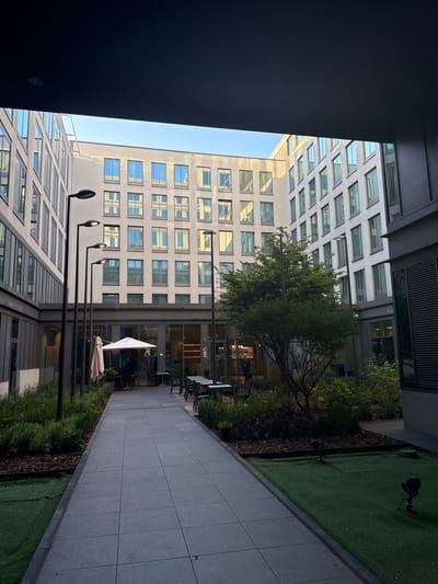 This image depicts a modern, well-maintained outdoor courtyard within a building complex, located in Saint-Gilles - Sint-Gillis, Belgium. The perspective is from beneath a dark, overhead architectural element, looking into the open central space.
The courtyard is surrounded by multi-story buildings featuring light-toned facades and numerous large, rectangular windows that reflect the sky and surrounding structures. A central pathway, constructed of grey square paving tiles, extends from the foreground towards the rear building.
To the left of the pathway, a landscaped area contains low green shrubs and mulched beds, interspersed with a line of four black, contemporary streetlights of varying heights. Further along this side, an outdoor seating area is visible, comprising multiple black tables and chairs sheltered by a large, open white patio umbrella, situated in front of a glass-walled section of a building.
To the right of the pathway, a larger landscaped area hosts a prominent, medium-sized deciduous tree with lush green foliage, alongside smaller shrubs and groundcover. In the immediate foreground on the right, a section of artificial green grass is visible, featuring two black, adjustable outdoor spotlights.
Through the glass facade of the rear building, one individual in dark clothing is observed standing inside, appearing to be in a reception or lounge area. No other persons are visible in the outdoor courtyard or seating areas. The overhead sky is clear blue, indicating daytime conditions, with soft shadows present within the courtyard.
