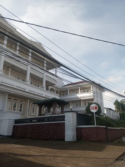The image shows a multi-story white building with columns and balconies. In front of the building is a decorative wall with a red brick pattern and a white pillar. An "OUT" sign with a red arrow is visible. Overhead, numerous power lines crisscross the sky. The scene appears to be a street view. The location in Phou Khoun, Laos, is not evident.