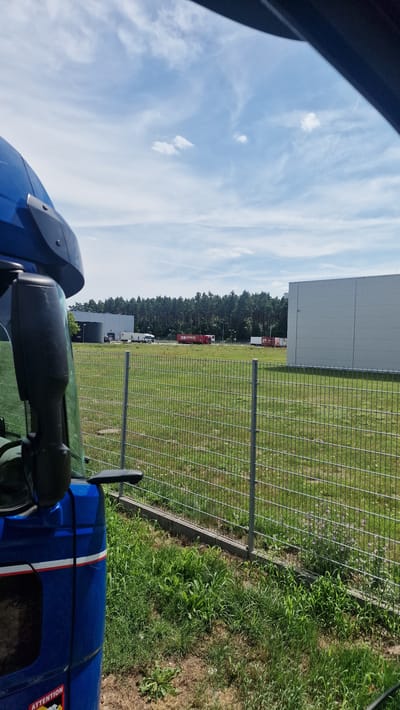 The image captures a view from inside a blue truck, looking out towards a fenced-off industrial area under a cloudy sky. The foreground features the side of the truck, including a side mirror. A metal fence with wire mesh runs horizontally across the mid-ground, separating the viewer from a grassy field. Beyond the field, several white and red trucks are parked or moving near large, modern buildings. A dense line of trees forms the backdrop. The location is identified as Stary Gostków, Poland. The scene appears to be a logistics or distribution center. There are no people visible in the image.