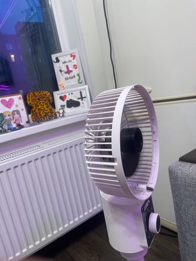 A white electric fan with a purple tinted grille is positioned on a wooden floor in front of a white radiator. The fan has a circular dial with numbers 0, 1, 2, and 3, and the brand name "LEBEN" is visible above the dial. To the left of the fan, on a white shelf, are several framed pictures and a stuffed animal with a leopard print pattern. The framed pictures contain various drawings, including a heart, stick figures, and what appear to be medical symbols. In the background, a window reflects a purple light from what looks like decorative string lights. The overall environment appears to be an indoor setting, possibly a room in a residential building in Shcheglovo, Russia.