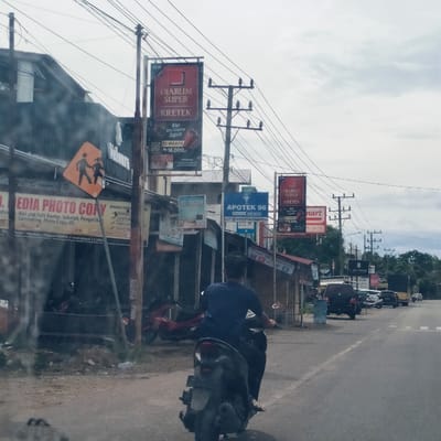 This street scene in Dayah Caleue, Indonesia, captures a busy commercial area under an overcast sky during daylight. The foreground shows a person from behind, riding a dark scooter motorcycle away from the camera. The street, marked with white lines, extends into the distance with several vehicles, including cars and a truck, either parked or driving.

On the left, a row of businesses with corrugated roofs lines the road. A prominent orange diamond-shaped sign indicating a pedestrian crossing is visible. Utility poles heavily laden with electrical wires stretch across the scene.

Visible text on signs indicates various businesses:
*   Multiple banners advertise "DJARUM SUPER KRETEK," stating "12 WRAPS Rp 18.000,-" and "KRETEKNYA SUPER".
*   "UD. MEDIA PHOTO COPY" offers "Alat alat Tulis Kantor, Sekolah, Pengetik, Laminating, Photo Copy, dll." and "MENERIMA PEMBAY[ARAN]" for "Listrik Praba, Speedy dan, Pusa All Op, Iren," with a location cue "Aceh - Medan (Depan SMPN 2 Indrajaya) Caleu."
*   A blue sign displays "APOTEK 96."
*   Parts of other signs are visible, including "mart" (likely a minimart), "atm," "SIG PONSEL," and "TOR RESMI VIVO" (a mobile phone outlet).
*   A partially visible word "Veloura" is seen on a building.

The scene depicts a typical everyday street with local commerce and active traffic.