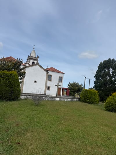 The image shows a small, white chapel in Castêlo da Maia, Portugal. The chapel has a red tile roof, a small bell tower, and a cross. It is surrounded by green grass and trees. A power pole is visible near the trees, on the right side of the chapel. The sky is blue with some clouds.