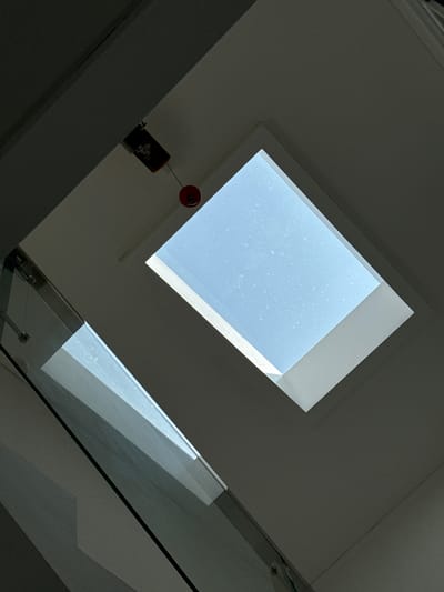 The image shows an interior view looking upwards towards a skylight. The skylight is rectangular and angled, revealing a light blue sky with small white speckles, possibly dust or condensation on the glass. Below the skylight, a dark ceiling angles downwards, creating a stark contrast with the illuminated sky.

In the lower left of the frame, a glass railing of a staircase is visible, reflecting the light from the skylight and the surrounding interior. The glass has a metallic frame. A dark, possibly wooden, beam runs diagonally across the upper left portion of the image, obscuring part of the ceiling. Attached to this beam, and hanging down, is a small, dark rectangular object with some orange and white markings, and below it, a small red round object attached by a thin wire.

The image does not contain any discernible elements that would specifically place it in Tanjungmeru, Indonesia, or any other city. The scene appears to be a modern interior space, possibly a home or a commercial building. There are no people, actions, or interactions depicted.