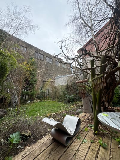The image displays an outdoor garden or courtyard scene located in Romainville, France, under an overcast sky. In the immediate foreground, a curvilinear lounge chair or sculpture, constructed from bent sheets of light-colored metal, rests on wooden decking. To its right, a segment of a round, slatted light-colored outdoor table is visible, with a green clip positioned on its surface. A set of bamboo-like wind chimes hangs from a multi-post wooden structure, which is intertwined with bare, gnarled branches, on the far right. A light-colored rectangular bin is partially obscured behind this structure.

The midground features a patchy grass lawn surrounded by various dormant deciduous plants, shrubs, and trees with bare branches, along with some evergreen foliage and a small tree exhibiting yellow blossoms. Disturbed earth with sparse vegetation borders the lawn. A silver-colored ladder is propped against an unseen structure on the far left.

In the background, a large, multi-story building constructed of dark stone or concrete dominates the left side of the frame. Its upper facade is extensively covered with bare climbing vines, revealing a row of rectangular, multi-paned windows. A portion of another building with a reddish-brown facade and a sloped roof is visible on the upper right, partially obscured by trees. No individuals are present in the scene.