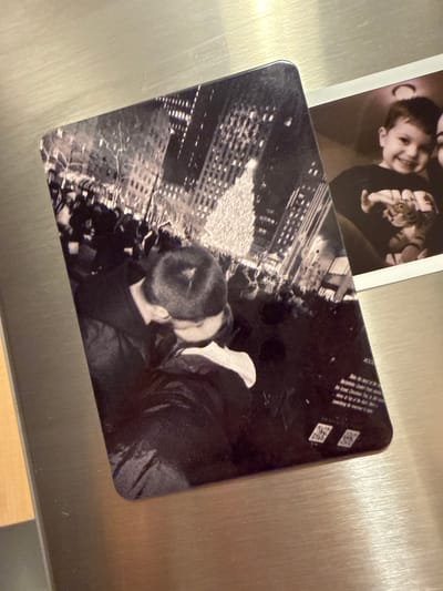 The image displays two photographs on a metallic surface, likely a refrigerator. The larger photograph, on the left, is a black and white image capturing a crowd gathered in front of a brightly lit Christmas tree, with tall buildings in the background. In the foreground of this photograph, a couple is embracing and kissing. The smaller photograph, partially visible on the right, is a color image of a young child smiling and looking towards the camera. The child is wearing a black shirt with a graphic design. The metallic surface shows a brushed texture. The overall scene appears to be an indoor setting, with the photographs mounted on a kitchen appliance. There is no discernible information in the image to indicate the location as the City of Yonkers, United States of America.