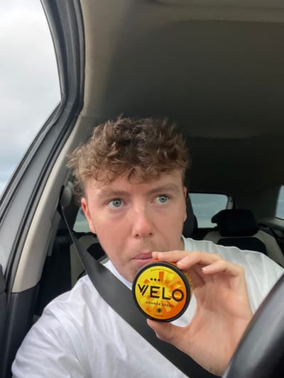 A single male individual is depicted seated in the driver's seat of a vehicle. He possesses light, curly hair and blue eyes, and is wearing a white t-shirt with a black seatbelt secured across his chest. His right hand holds a circular, orange and yellow tin labeled "VELO ORANGE SPARK" with "NICOTINE POUCHES" visible at the top edge. Four black dots are present above the "VELO" branding. The subject's gaze is directed slightly away from the camera, towards the upper right of the frame. The interior environment shows a dark-colored car door panel and A-pillar on the left, with an overcast sky visible through the driver's side window. Portions of the steering wheel and rear passenger seats are also discernible. The scene is situated in Barmston, United Kingdom.