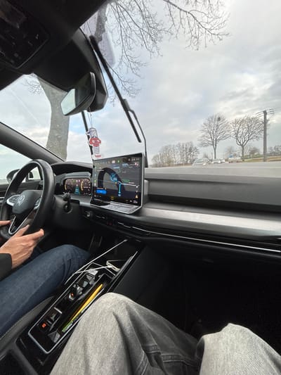 The image depicts the interior of a modern Volkswagen vehicle from the front passenger perspective, looking forward through the windshield. The scene is located in La Wantzenau, France, with the GPS navigation screen displaying a route on "Rue des Berges" in this city, indicating the time as 13:25 and an ambient temperature of 22.0°C.
A driver, wearing blue jeans and a dark top, is seated on the left, with both hands on the steering wheel. The front passenger's legs, clad in grey jeans, are visible on the right. The dashboard features a large central infotainment screen and a digital instrument cluster. A red and white spring-toy air freshener hangs from the rearview mirror. The center console contains charging cables, a smartphone, and a yellow writing utensil in various storage compartments. The windshield shows water droplets, and the wipers are visible, suggesting recent or ongoing precipitation.
Outside the vehicle, the environment features a paved road with barren deciduous trees lining both sides, indicative of winter or early spring. A distant treeline is visible under an overcast, light grey sky. Several vehicles, including cars and a white truck/van, are present on the road in the distance. A utility pole with power lines is visible on the right side of the road.