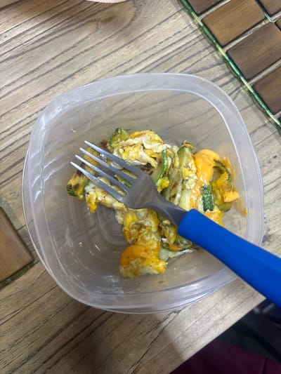 A close-up, high-angle shot shows a clear plastic food container filled with a mixture of scrambled eggs, sliced zucchini, and a creamy orange sauce, possibly cheese sauce. A blue-handled fork is inserted into the food. The container sits on a table or surface with a wood-grain pattern, alongside a section of a decorative table with a mosaic of small wooden tiles. There is no indication of the city of Buenavista, Mexico.