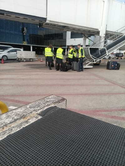 This image depicts an airport tarmac scene at Gran Canaria Airport in Ojos de Garza, Spain. In the foreground, the edge of a weathered, light-colored metal surface with peeling paint and rust is visible on the left, leading to a dark grey, textured mat with a grid pattern covering the bottom right.
In the midground, a group of approximately seven individuals is gathered on the concrete tarmac, near the base of a white jet bridge. Most individuals, including at least two men and five women, are dressed in dark uniforms augmented by bright fluorescent yellow-green safety vests with dark lettering on the back. Several pieces of rolling luggage, primarily black and grey suitcases, are positioned on the ground around the group. One female individual is observed looking at a mobile phone.
To the left of the group, a white car, possibly a station wagon, is parked. Adjacent to it is a rectangular piece of white ground support equipment on wheels and an orange traffic cone. The tarmac is light grey with distinct red and white painted lines.
In the background, a large white jet bridge structure extends towards a terminal building. A set of stairs descends from the jet bridge to the tarmac on the right. Behind the jet bridge, a modern airport terminal building with large, dark blue reflective glass windows is visible. The sky above is bright and overcast.