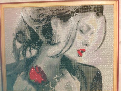 This is a diamond painting featuring a stylized portrait of a woman. The woman is depicted in grayscale with a bright red element, possibly a rose or a flower, held in her hand or near her chest. Her hair is dark and appears to be styled in waves. Her face is partially obscured, with one side appearing more in focus than the other. There is a hint of another face or figure in the background, also in grayscale. The red of the flower is a stark contrast to the otherwise monochromatic image, drawing the viewer's eye. The background is a textured pattern of light and dark squares, characteristic of diamond art. The artwork is framed in a wooden frame with a light-colored matting. The overall impression is one of artistic expression, focusing on contrast and form. There is no visible text in the image. The setting is abstract, with no discernible location cues. The emotion conveyed is ambiguous, possibly contemplative or dramatic due to the contrast and stylized depiction.