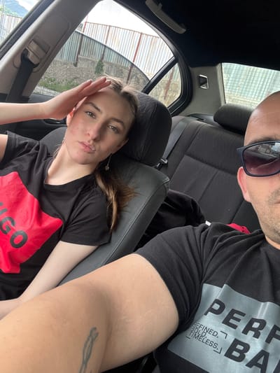 In Vlahovo, Bulgaria, a man and a woman are photographed inside a car, likely a sedan, with the man in the foreground and the woman to his left. The man is wearing a black t-shirt with a silver graphic that reads "PERF" and "BA." He is also wearing dark sunglasses, and his head and shoulder are visible. The woman is wearing a black t-shirt with a large red graphic that appears to say "HUGO." She is resting her hand on her forehead and looking at the camera. The interior of the car is visible, including the seats and seatbelts. The background outside the car shows a fence and some greenery, suggesting an outdoor setting in a rural or semi-rural area.