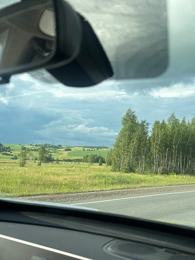 The image depicts a road trip scene viewed from inside a vehicle. The perspective is from the passenger seat, looking outwards and slightly upwards. The main subject is a landscape featuring a road on the right side, bordered by a grassy verge and a line of trees. Beyond the trees, rolling green fields are visible, with power lines stretching across the horizon in the distance. The sky is partly cloudy, with a mix of bright and dark clouds, suggesting variable weather. A portion of the car's interior, including a rearview mirror and a sun visor, is visible in the foreground on the left, casting shadows on the windshield. The overall impression is that of a journey through a rural or semi-rural area under a dynamic sky. The time of day appears to be late morning or early afternoon, given the lighting conditions. There is no visible text in the image. The location is identified as Bekhterevo, Russia, which implies a likely Eastern European or Russian landscape.