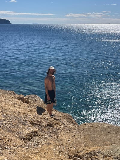 An adult male subject is positioned on a rugged, light-brown rocky cliff overlooking a vast expanse of water under a clear blue sky with sparse clouds, in Megalochori, Greece. The subject is bare-chested, displaying visible chest hair and tattoos on his left arm and shoulder area. He wears a light-colored baseball cap, dark shorts with a white graphic design on the left leg, and dark closed-toe footwear. His dark, wavy hair extends from beneath the cap. He holds a metallic, silver-colored bottle in his right hand. The subject is facing the viewer, smiling. The water, identified as the sea, exhibits a deep blue hue with significant solar glinting creating shimmering patterns across its surface, especially on the right side of the frame. A distinct, dark landmass is visible on the distant left horizon, with additional fainter shapes further along the horizon. Direct sunlight casts a short shadow behind the subject on the uneven, rocky terrain.