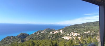 This panoramic image captures a breathtaking coastal landscape in the Municipal Unit of Lefkada, Greece, viewed from a high vantage point. The scene is dominated by the vast, deep blue Ionian Sea on the left, contrasting with the lush green, tree-covered hillsides on the right.

The sky is a clear, vibrant blue, with a few delicate, wispy white clouds appearing towards the right horizon. The sea exhibits a striking color gradient, transitioning from a rich, dark blue in the distance to a brilliant, lighter turquoise closer to the shore. Faint outlines of distant landmasses are visible on the horizon across the sea.

The rolling hills are densely forested with dark green trees, likely pines and cypresses, creating a rich, natural tapestry. Numerous white and light-colored buildings, possibly homes or holiday accommodations, are nestled among the trees on the slopes, forming a scattered village or residential area. Some parts of the hillside show patches of exposed earth, suggesting ongoing development or construction.

The photograph was taken during bright daylight, under clear and sunny weather conditions. The composition is partially framed on the right and top by a dark, vertical and horizontal architectural element, likely a window or balcony frame, implying the photo was taken from an indoor space or a sheltered viewing platform. A subtle reflection of the top of a person's head or hair is visible at the very bottom edge, indicating an observer. There is no visible text in the image.