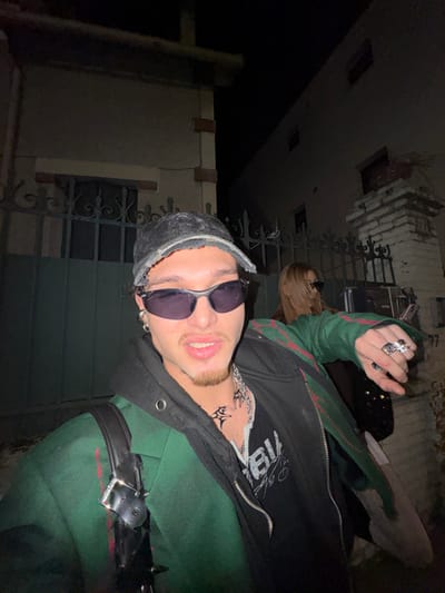 A night scene depicts two individuals outdoors in what appears to be an urban or suburban setting, specifically stated as Asnières-sur-Seine, France. The precise location within the city is not identifiable from visual cues.
In the foreground, a male individual is positioned centrally, facing the camera as if for a close-up photograph. He wears a distressed black baseball cap, dark wrap-around sunglasses, and multiple silver hoop and stud earrings. His face features a thin goatee and mustache, and his lips are slightly parted. Neck tattoos include a tribal-style design on the left side and a chain-link pattern on the right. He is dressed in a layered ensemble consisting of a black hooded zipper jacket worn over a black t-shirt with partially visible white text, and an outer dark green jacket with red stripe accents on the sleeves and body. Accessories include a silver chain necklace, multiple silver rings on his right hand, and a black backpack with studded straps visible over his left shoulder. His right arm is bent, with his hand near chest level.
Behind and to the right of the foreground subject, a second individual, appearing female, is partially visible. She has long, light brown hair and wears dark sunglasses. Her attire includes a dark green jacket with red accents, mirroring the style of the foreground subject's outer jacket. She faces away from the camera, looking towards the right side of the frame. Her left hand is holding a dark, possibly textured or sequined, bag.
The background consists of architectural structures and a fence, illuminated by what appears to be a camera flash. To the left, a light-colored building with dark brick-like accents at the corners and around a window is visible. To the right, a darker, multi-story building is present. A dark, ornate metal fence or gate runs horizontally across the midground, situated between the subjects and the buildings. The overall environment is dimly lit under a dark sky, suggesting nighttime. Sparse, dry vegetation is visible near the base of the rightmost building.