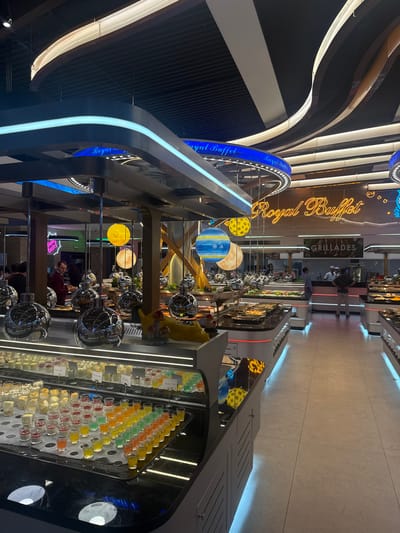 The image shows the interior of a "Royal Buffet" restaurant. There's a contemporary design with curved, illuminated architectural elements. Various food stations are visible, showcasing desserts, salads, and cooked dishes. Some stations have spherical decorations. A "Grillades" sign is also present, and customers can be seen interacting with the buffet offerings.