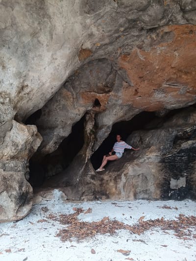A person is sitting inside a cave in Thap Put, Thailand. The cave has multiple openings and is composed of gray and brown rock. The person is wearing a striped shirt, denim shorts, and sandals. The floor of the cave and the area outside are covered in white sand mixed with brown leaves.