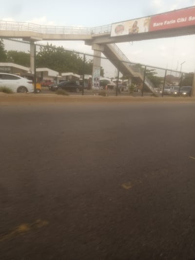 The image shows a pedestrian bridge over a street in Unguwan Dosa, Nigeria. The bridge is made of concrete and has metal railings. There is a large advertisement banner on the side of the bridge that reads "Bare Farin Ciki Sa". Below the bridge, there are several vehicles on the street, including cars, vans, and a red utility vehicle. A white SUV is parked on the left side of the image, partially obscured by a fence. A black car is driving on the street, and several other vehicles are visible in the background, including white vans.

There are some people visible near the street level, some of whom appear to be vendors or pedestrians. The scene suggests a typical urban environment with traffic and commercial activity. The sky is overcast, indicating a cloudy day. The time of day appears to be late afternoon or early evening, based on the lighting conditions. The overall atmosphere is one of a busy public space. There are also some trees and buildings visible in the background, contributing to the urban landscape.