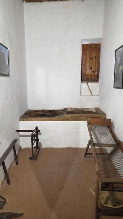 The image depicts the interior of a room, likely within a historical building, potentially in the city of Altura, Spain. The room features white walls and a terracotta-colored floor.

On the left, the white wall is punctuated by a small, rectangular wooden window or shutter. Above, a framed picture hangs on the wall.

Along the wall towards the right, there is a series of architectural features that appear to be integrated into the wall structure. These include an arched alcove, a circular opening, and a rectangular opening, all possibly remnants of former doorways or functional spaces.

On the floor, a metal rack or stand is positioned. Above it, a wooden cage or enclosure is present, containing hay.