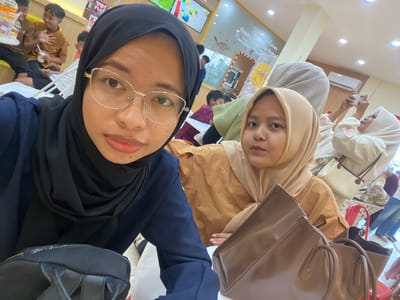 The image appears to be a selfie taken indoors, possibly at a cafe or bakery named "Baking Room". The foreground is dominated by two young women wearing hijabs. The woman on the left is wearing a black hijab and a dark blue top, with gold-rimmed glasses. She is holding a black bag. The woman on the right is wearing a light brown hijab and a tan top. She is holding a brown handbag. In the background, several children and adults are visible. Some children are sitting at a table, and one appears to be holding a cup. Another woman in the background is taking a photo with her phone. The environment suggests a casual dining or social setting with light-colored walls and artificial lighting. There are signs and decor visible on the walls, including one that says "Baking Room". Based on the context of "Kembang Harum, Indonesia", it is likely that this establishment is located in that area.