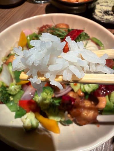 A close-up shot shows a pair of wooden chopsticks holding a portion of white rice. The background features a salad with ingredients like broccoli, red peppers, red onions, yellow peppers, and glazed nuts or meat, presented in a white bowl. Other bowls are partially visible on a wooden surface. The location information (Kiryat Ono, Israel) is given, but not directly evident from the image.