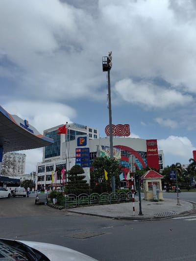 The image depicts a bustling urban street scene in Thuận Hóa Ward, Vietnam, under a partly cloudy sky. In the foreground on the left, a "Petrolimex" gas station canopy is visible, with two white vehicles and a person in a light blue shirt near the fuel pumps. Adjacent to the gas station is a multi-story building featuring blue-tinted windows, a "P" logo, and a Vietnamese flag flying on a pole. An electronic display board shows fuel prices. A small, white utility cart is parked on the curb nearby.
In the midground, a large commercial building, identified by a prominent red "GO!" logo at its peak, dominates the right side of the frame. Below the main logo, "Kohn" and "LOTTERIA" signs are visible. This building has a modern, multi-colored facade. A tall, grey utility pole with an array of fixtures at its summit stands between the gas station and the "GO!" building.
At a street corner in the center-right, a small, red-roofed kiosk or booth is situated on a paved island, next to a red and white striped traffic light pole. The area also features a manicured green space with trimmed hedges and small trees, bordered by a green metal fence. The street is paved, and multiple motorcycles, some with riders, are visible on the road, indicating active traffic. Palm trees and other buildings are present in the background.