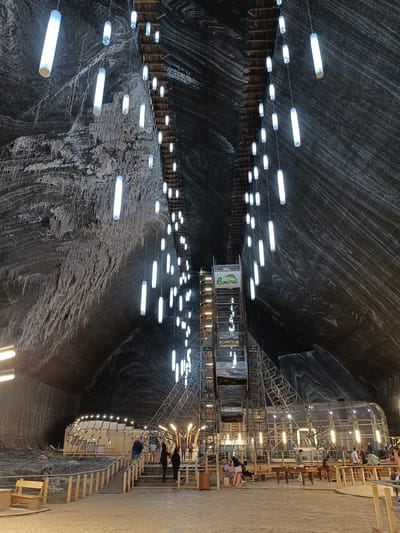 The image captures an interior view of a salt mine attraction, likely a tourist destination in Turda, Romania. The setting is a vast cavernous space with rough, textured walls that display swirling patterns of dark and light salt deposits. Hanging from the ceiling by thin wires are numerous cylindrical, white fluorescent lights, creating an ethereal glow. In the center of the cavern, a large, multi-level metal structure, resembling a scaffold or an elevator shaft, rises towards the ceiling. A sign on this structure reads "România" with a green logo featuring hills and a sun.
Below, a sandy floor is dotted with wooden benches and tables, suggesting an area for visitors to rest or engage in activities. Several people are visible walking around, sitting on benches, and interacting with each other, indicating that the location is open to the public. Some individuals are gathered near stairs leading up to the central structure, while others are seated at tables. The lighting suggests it is either evening or the cavern is illuminated artificially. The overall atmosphere appears calm and recreational, a stark contrast to the geological formation of the salt mine. The texture of the salt walls, the hanging lights, and the man-made structures combine to create a unique and intriguing visual experience.