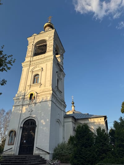 An exterior, low-angle, full shot captures the white brick bell tower of a Russian Orthodox church in Zagornovo, Russia. The bell tower, ornate with decorative brickwork, features arched openings where bells hang and is topped with a golden dome and cross. A smaller dome and cross crown the main church building, visible to the right of the bell tower. The building is surrounded by evergreen trees and a clear blue sky. Sunlight casts shadows on the tower, indicating it is late afternoon. The entrance to the bell tower is a large, dark wooden double door, accessed by a set of stone stairs. An icon is mounted on the left side of the tower above the entrance.