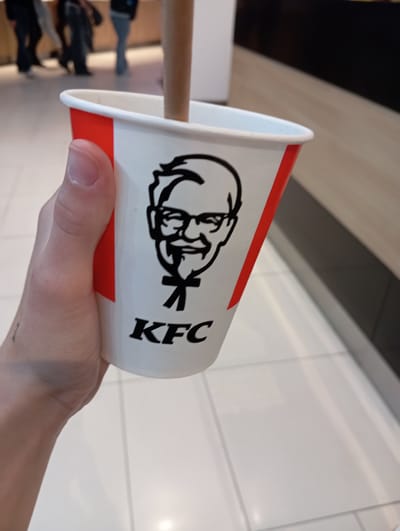 The image shows a hand holding a KFC branded cup, with a straw inside. The cup is white with red accents and features the Colonel Sanders logo. In the background, blurry figures of people are walking in what appears to be a public indoor space, possibly a shopping mall or food court. The floor is tiled in a light color. The environment suggests a commercial setting within the city of Wołomin, Poland, where KFC establishments are present.