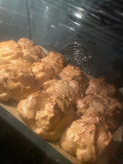 A batch of golden-brown pastries, likely profiteroles or cream puffs, are baking in an oven. They are arranged on a parchment-lined baking sheet, puffed up, and slightly uneven in shape. The interior of the oven is visible, including the heating element fan. Steam or slight condensation is apparent inside the oven, contributing to a hazy view. The overall scene suggests the final stages of baking, with the pastries developing their characteristic crispy texture.