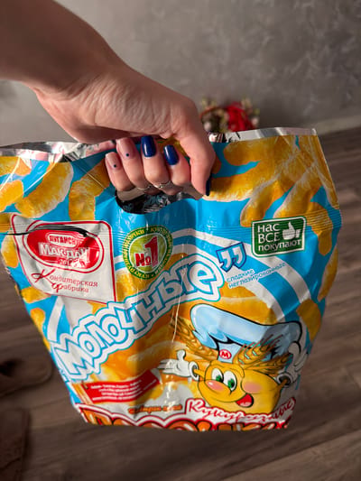 A person's hand, adorned with nude and navy blue nail polish, holds a large, crinkled plastic bag of "Molodnye" brand puffed corn snacks. The bag features a bright blue and yellow design with images of puffed corn and a cartoon character wearing a chef's hat. Text on the bag is in Russian and includes brand names and product descriptions. The background consists of a textured gray wall and a wooden floor. The image is cropped to focus on the bag and the hand. The location is not specified in the image.