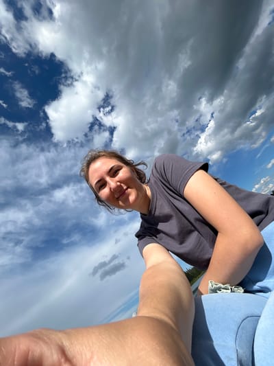 The media file is a selfie taken from a low angle, looking up at the sky. The main subject is a person, likely female, with brown hair, who is smiling. They are wearing a dark grey t-shirt and light blue shorts with a frayed hem. The background is dominated by a dramatic sky with a mix of dark, stormy clouds and bright blue sky with scattered white clouds. The lighting suggests it is daytime, possibly late afternoon or early evening, given the dynamic cloud formations. The perspective creates a sense of looking up from a ground-level position, possibly while lying down or sitting on the ground. The overall mood is casual and relaxed, with the subject appearing happy and enjoying the outdoors. The location context provided is Верх-Рождество, Russia. No visible text is present in the image.